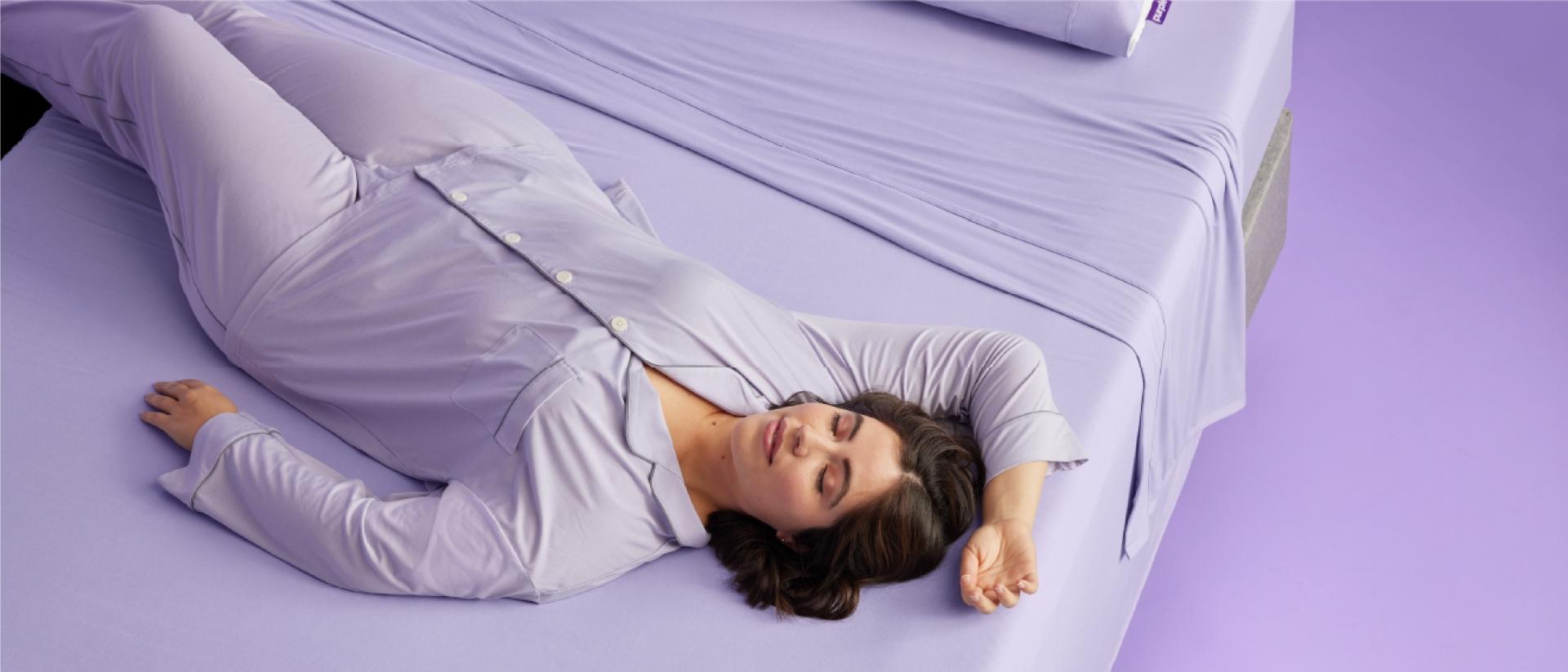 A woman in purple pajamas lying on a bed with purple sheets.