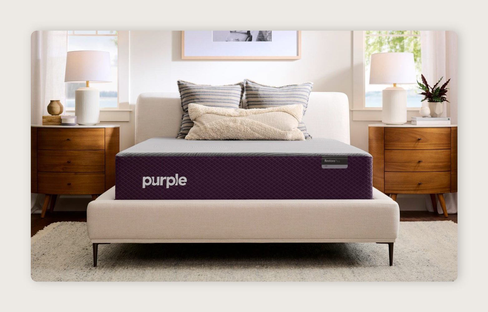 The Purple RestorePlus™ Hybrid Mattress on a cream upholstered bed frame between two wooden nightstands. 