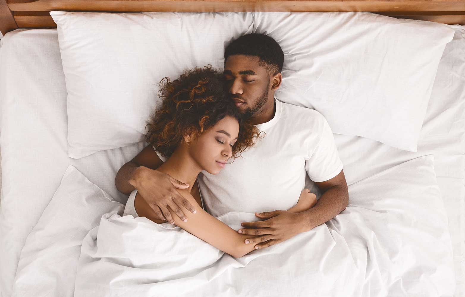 A couple cuddling together in a california king bed with white sheets and white pillows. 