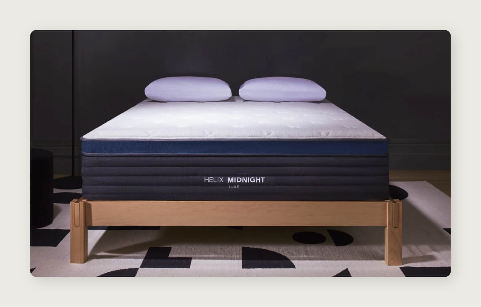 A Helix Midnight Luxe mattress with two pillows on a light wood platform bed frame in a dark room.