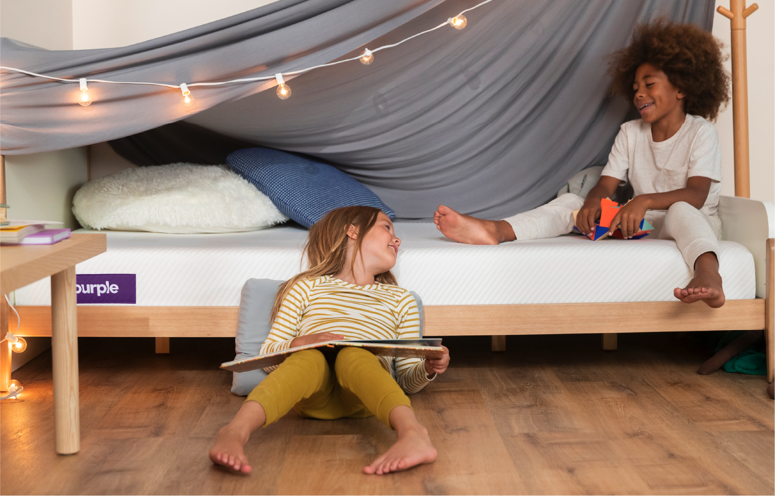 Two children playing in a bedroom with a Purple Kid’s Mattress, using a Purple kids’ sheet to make a blanket fort.