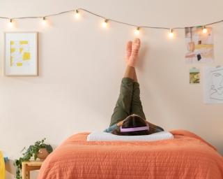 A girl wearing purple headphones lies on a full size bed on her back with her legs up the wall in a teen bedroom.