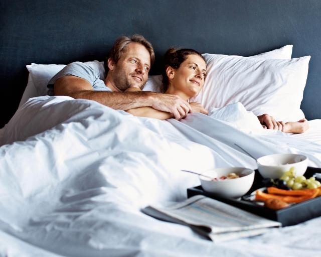 A man and woman lay in bed looking off to the side with breakfast on a tray sitting on the mattress..
