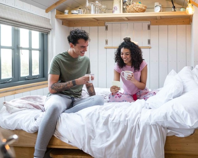 A young couple drinking coffee on a short queen mattress in their RV.