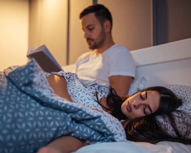 A photo of a woman sleeping in bed and her partner is sitting up to read. 