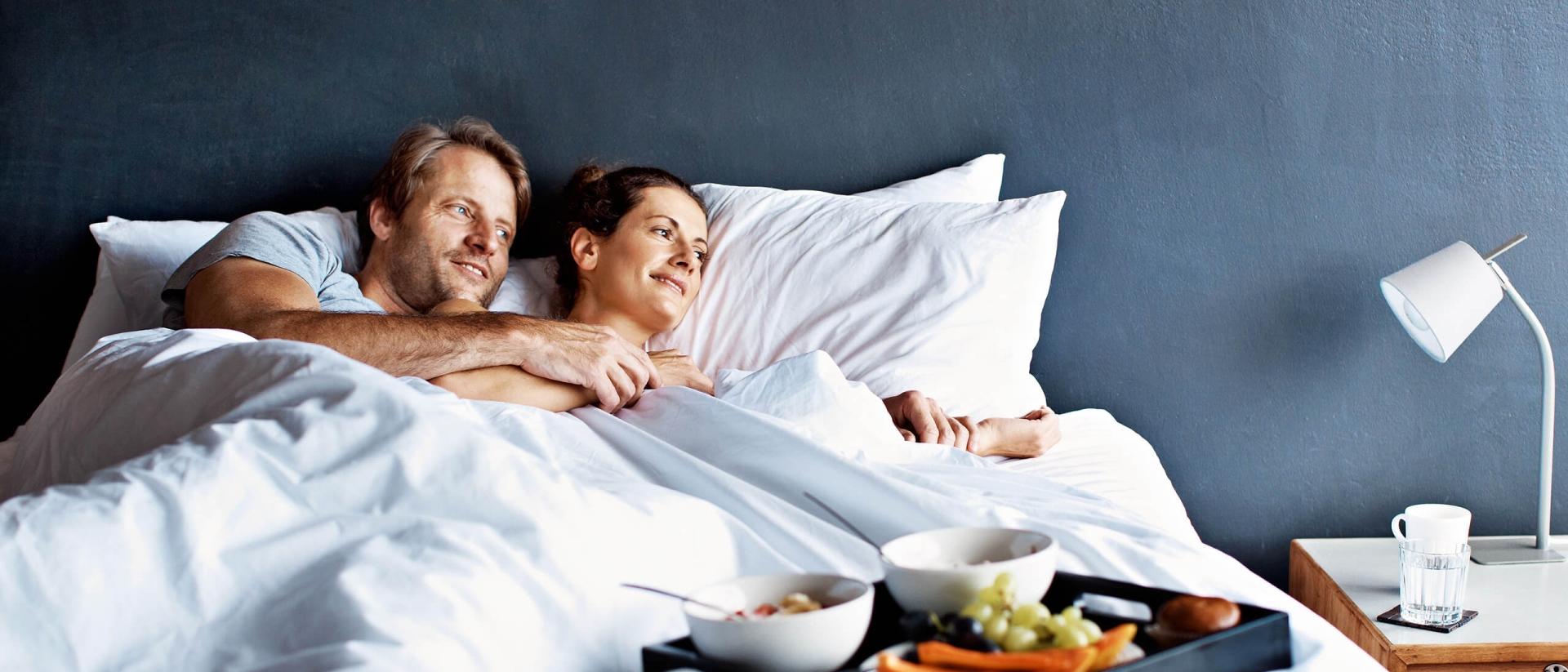 A man and woman lay in bed looking off to the side with breakfast on a tray sitting on the mattress..
