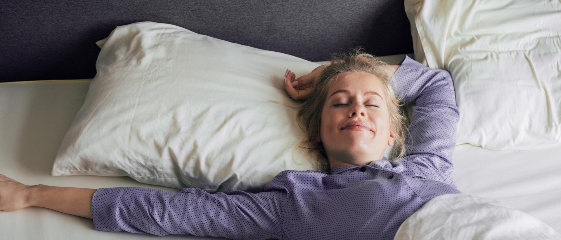 A woman wearing purple pajamas wakes up smiling in a bed with white sheets.