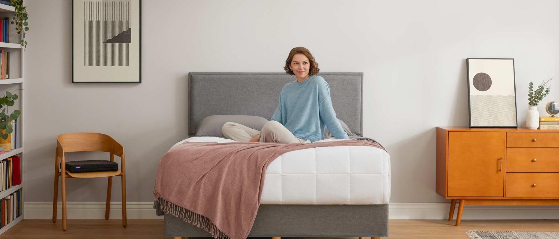 A woman sits upright on top of the covers of a bed on the Purple Bed Frame.