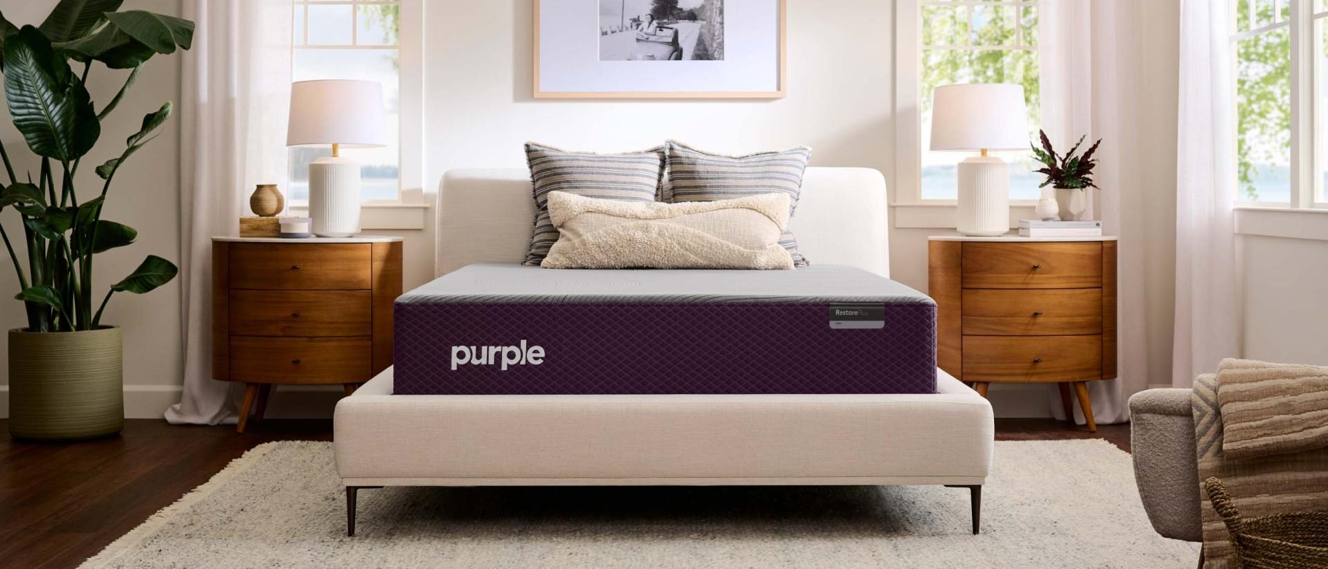 The Purple RestorePlus™ mattress on a cream upholstered bed frame between two wood nightstands in a bright bedroom.