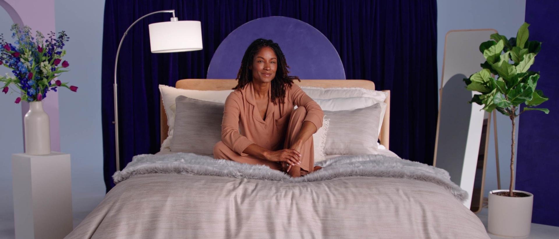 Woman in tan pajamas sits on a bed with gray bedding in a bedroom with blue walls and plants.
