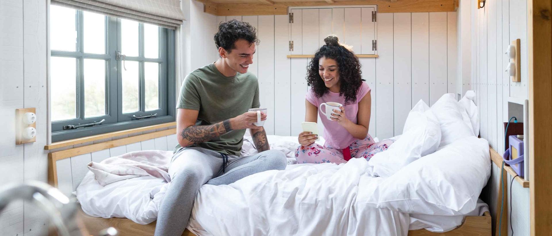 A young couple drinking coffee on a short queen mattress in their RV.