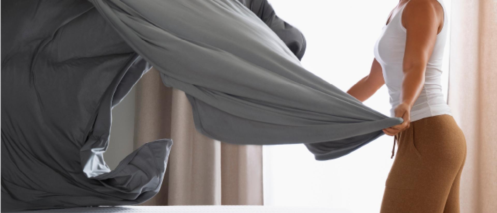 A woman fanning out sheets over a bed.